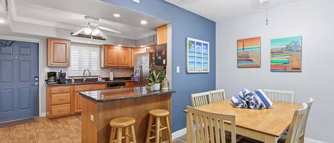 A modern kitchen with wooden cabinets, a central island with stools, a dining area with a wooden table and chairs, and artwork on the walls.