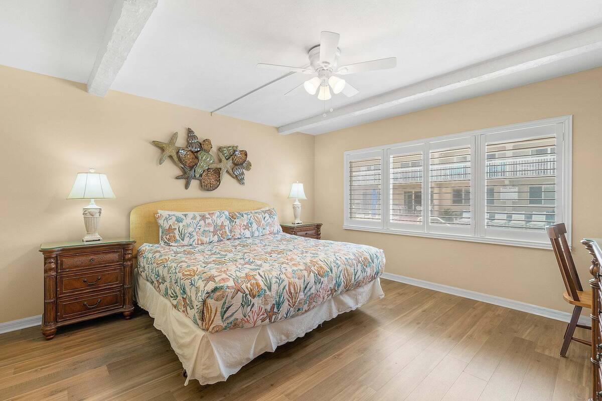 Une chambre à coucher comprenant un lit, des tables de chevet, un ventilateur de plafond, des éléments décoratifs sur les murs, une fenêtre avec des stores et une chaise.