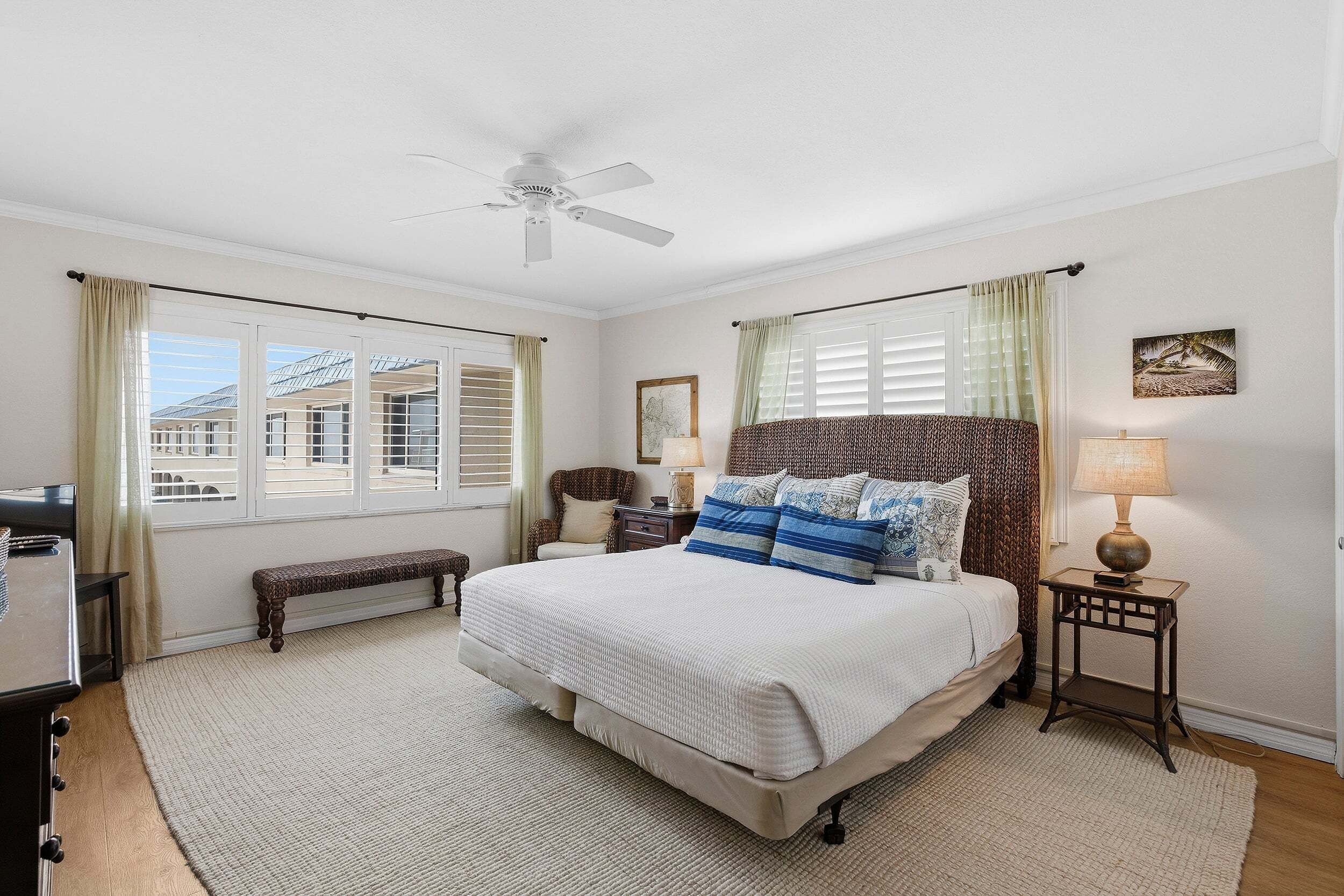 A bedroom with a bed, a bedside table, a lamp, a bench, a ceiling fan, and large windows.