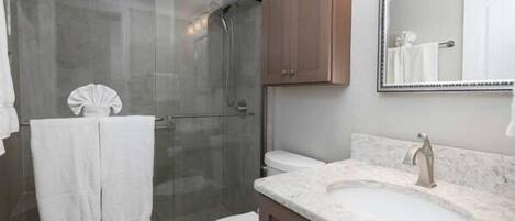 A bathroom with a glass shower enclosure, a white toilet, a marble sink, and wooden cabinets.