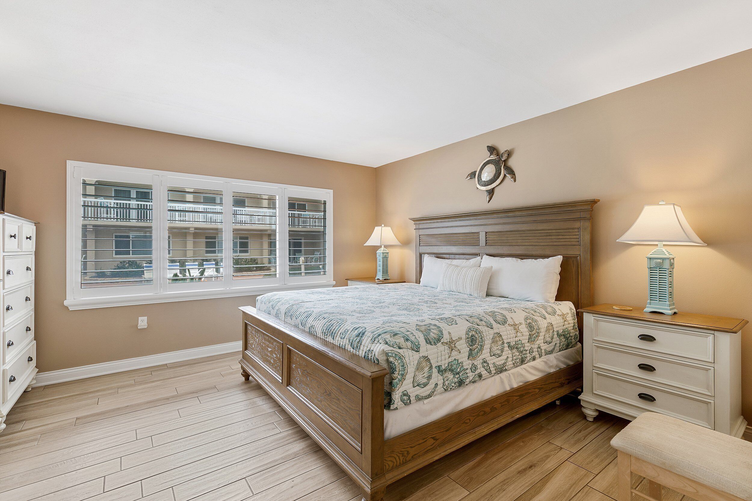 A bedroom with a large bed, a nightstand with two lamps, a dresser, and a window with a view of a balcony.