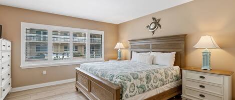 A bedroom with a large bed, a nightstand with two lamps, a dresser, and a window with a view of a balcony.