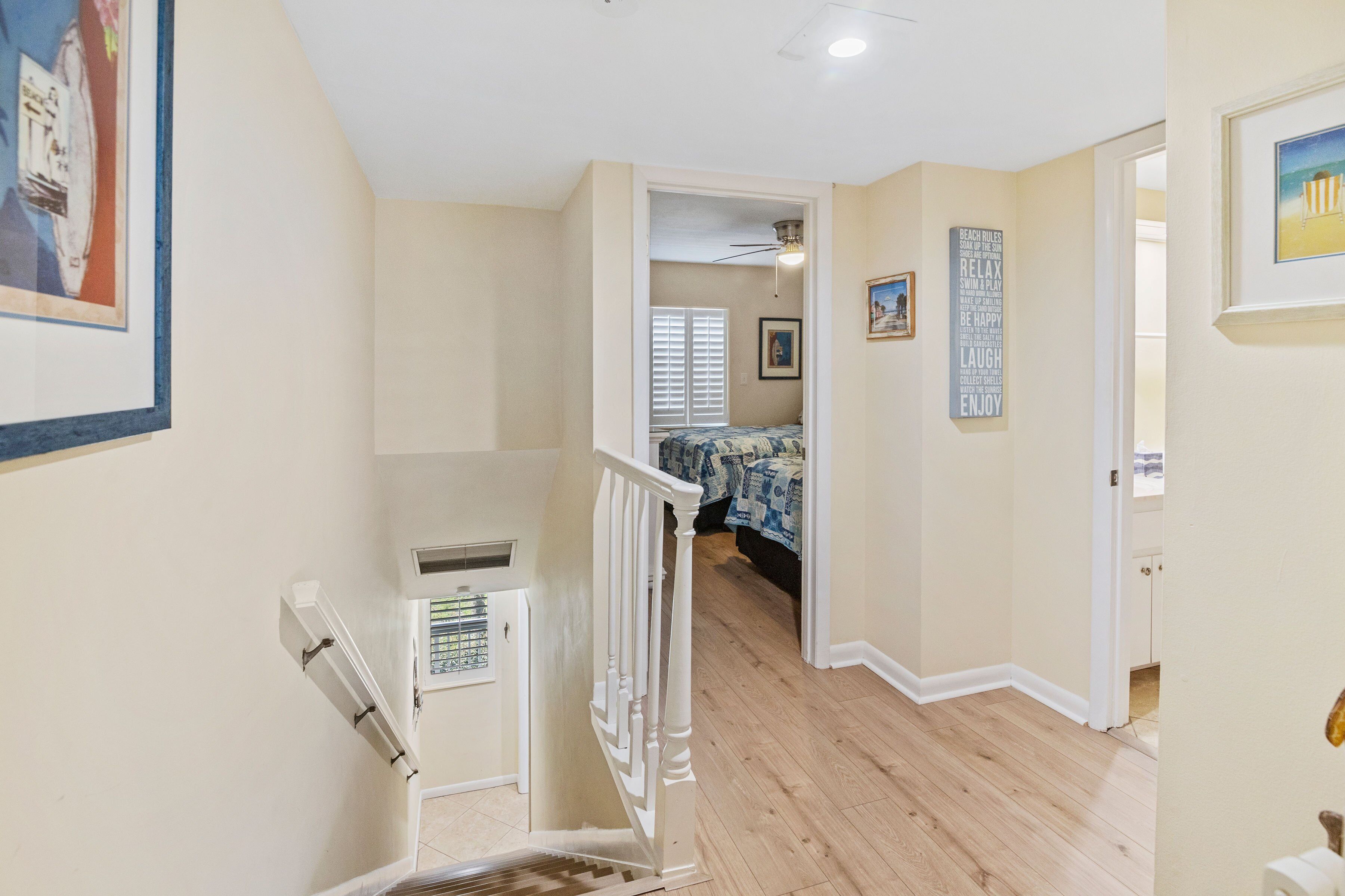 A hallway with a staircase leading to a bedroom with a bed and a ceiling fan. There are framed pictures on the walls and a door to another room.