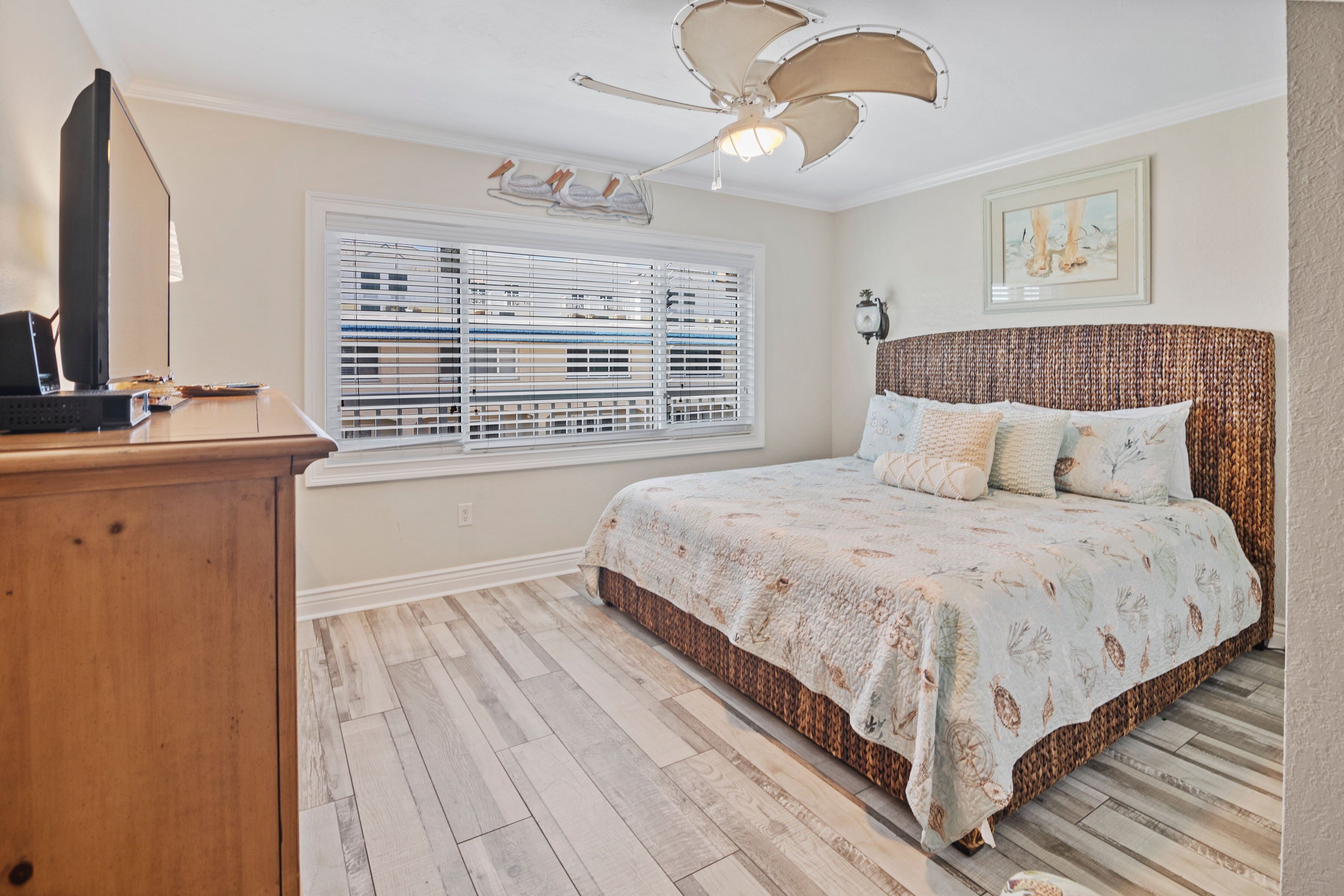 A bedroom with a bed, a desk, a ceiling fan, a window with blinds, and a wall-mounted light fixture.