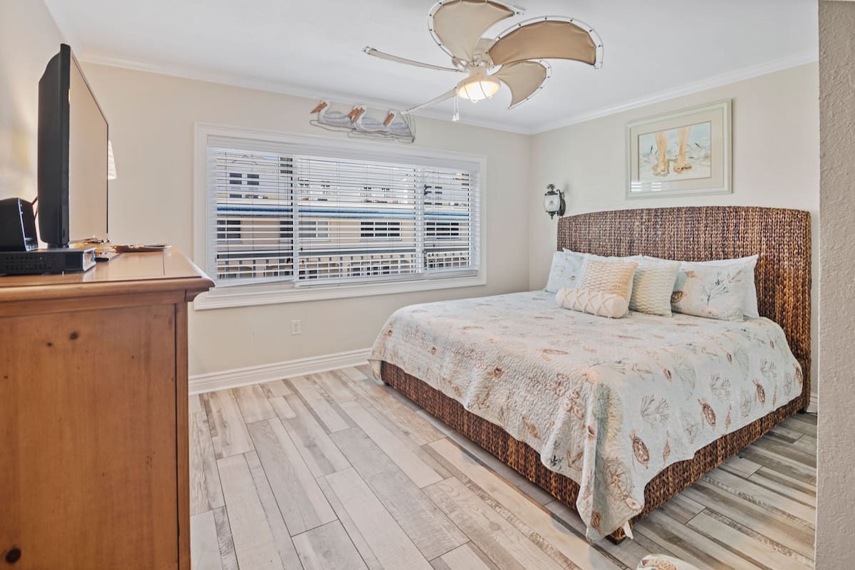 A bedroom with a bed, a desk, a ceiling fan, a window with blinds, and a wall-mounted light fixture.