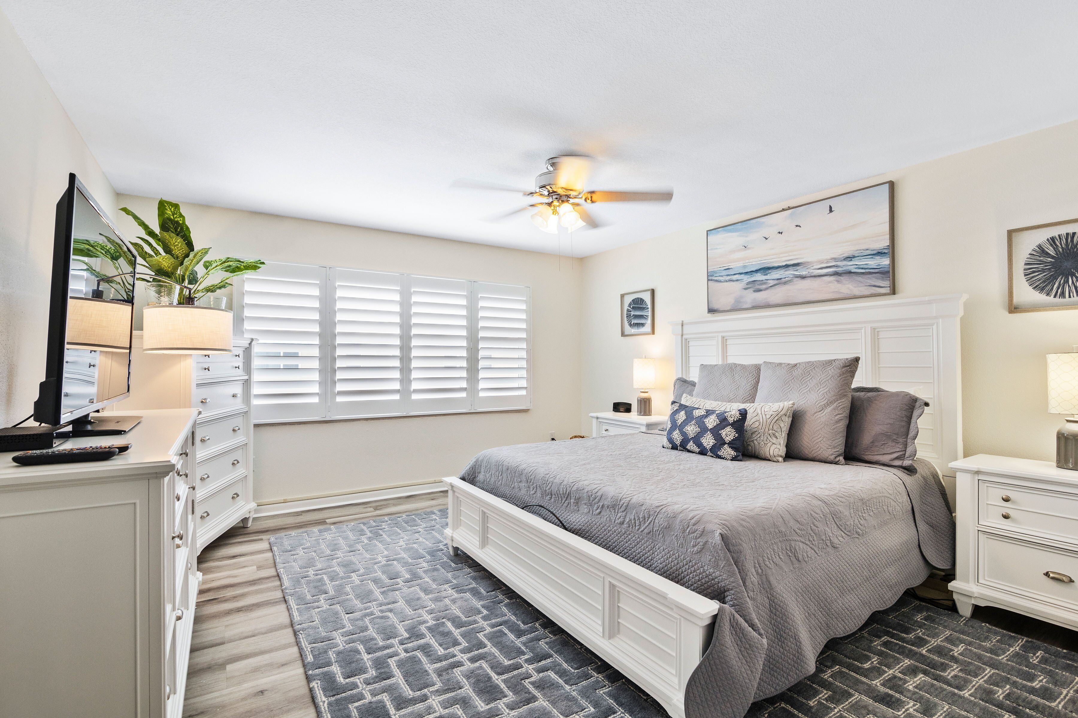 A bedroom with a bed, a ceiling fan, a window with blinds, a bedside table with a lamp, and a dresser.