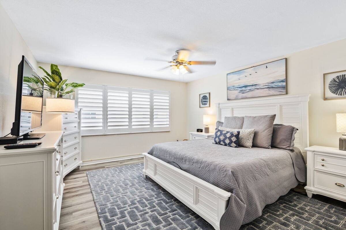 A bedroom with a bed, a ceiling fan, a window with blinds, a bedside table with a lamp, and a dresser.