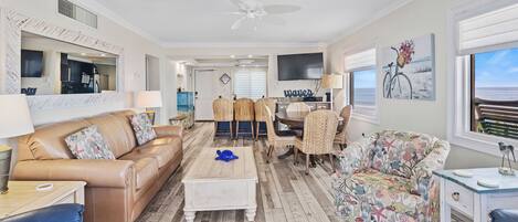 A spacious living room with a brown leather sofa, a coffee table, wicker chairs, and a patterned armchair. A television is mounted on the wall, and there is a large window with a view of the ocean.