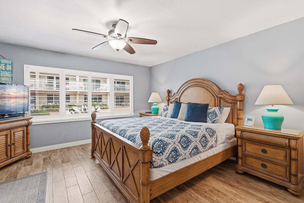 Une chambre à coucher comprenant un lit, des tables de chevet, un ventilateur de plafond, une fenêtre et une vue sur les bâtiments environnants.