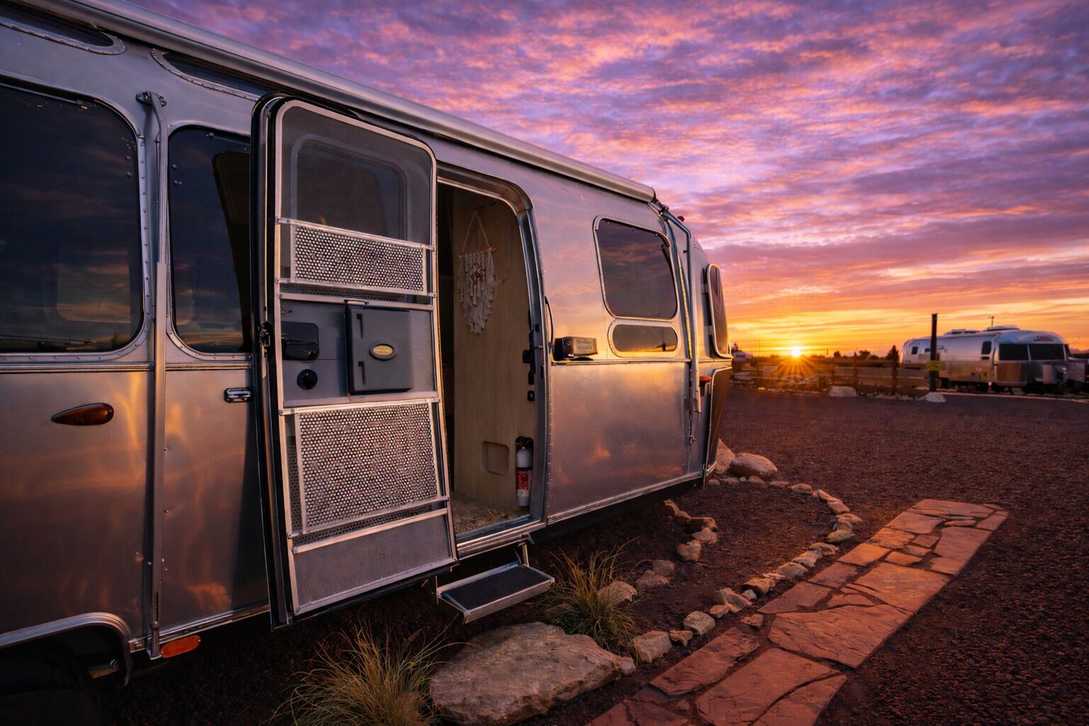 A silver RV with a sliding door, a small step leading up to the entrance, and a gravel driveway.