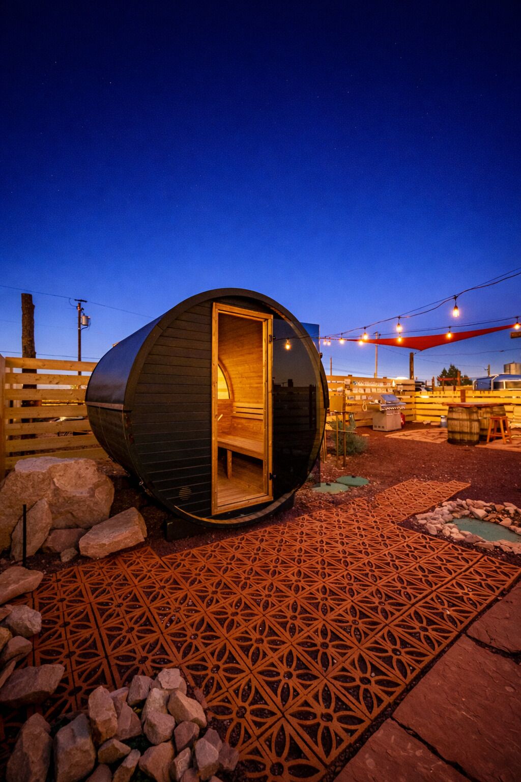 Une structure sphérique en bois, en forme de capsule, dotée d’une porte ouverte, située sur un sol pavé à motifs, avec des rochers et une clôture en bois en arrière-plan.