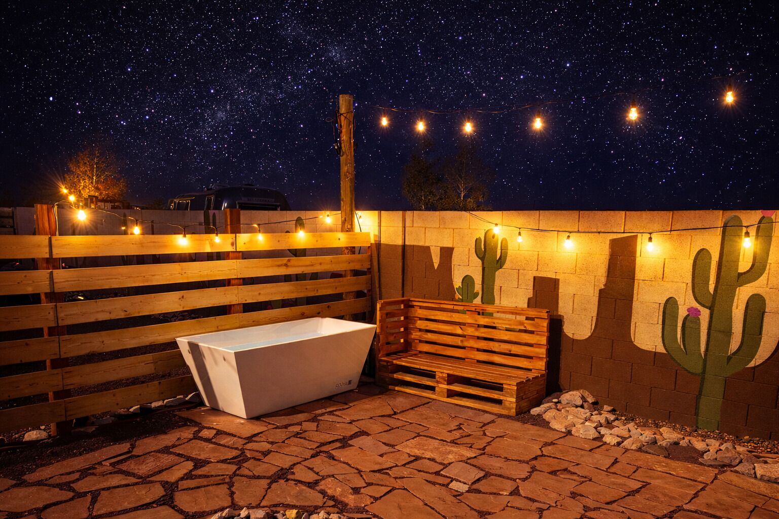 An outdoor patio with a wooden bench, a white bathtub, and a cactus mural under a starry night sky.