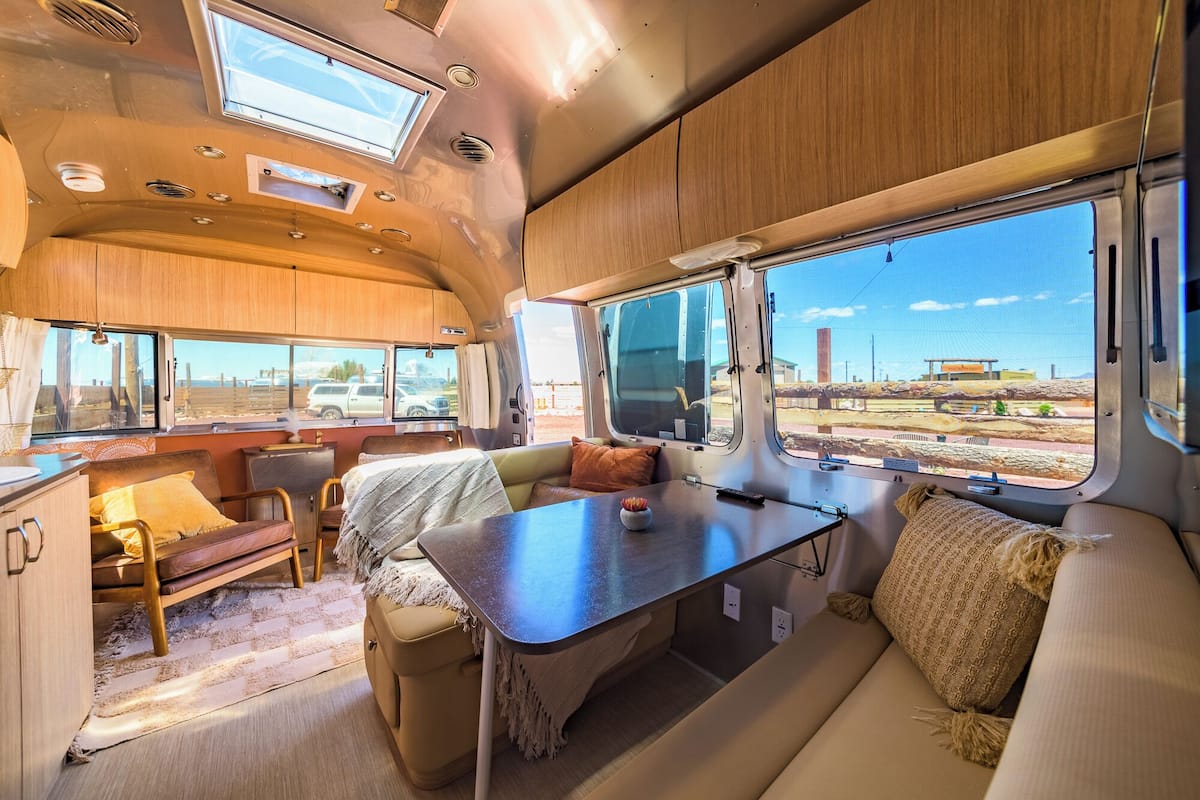 Interior luminoso de una caravana, con zona de comedor, asientos y amplios ventanales que ofrecen vistas al exterior.