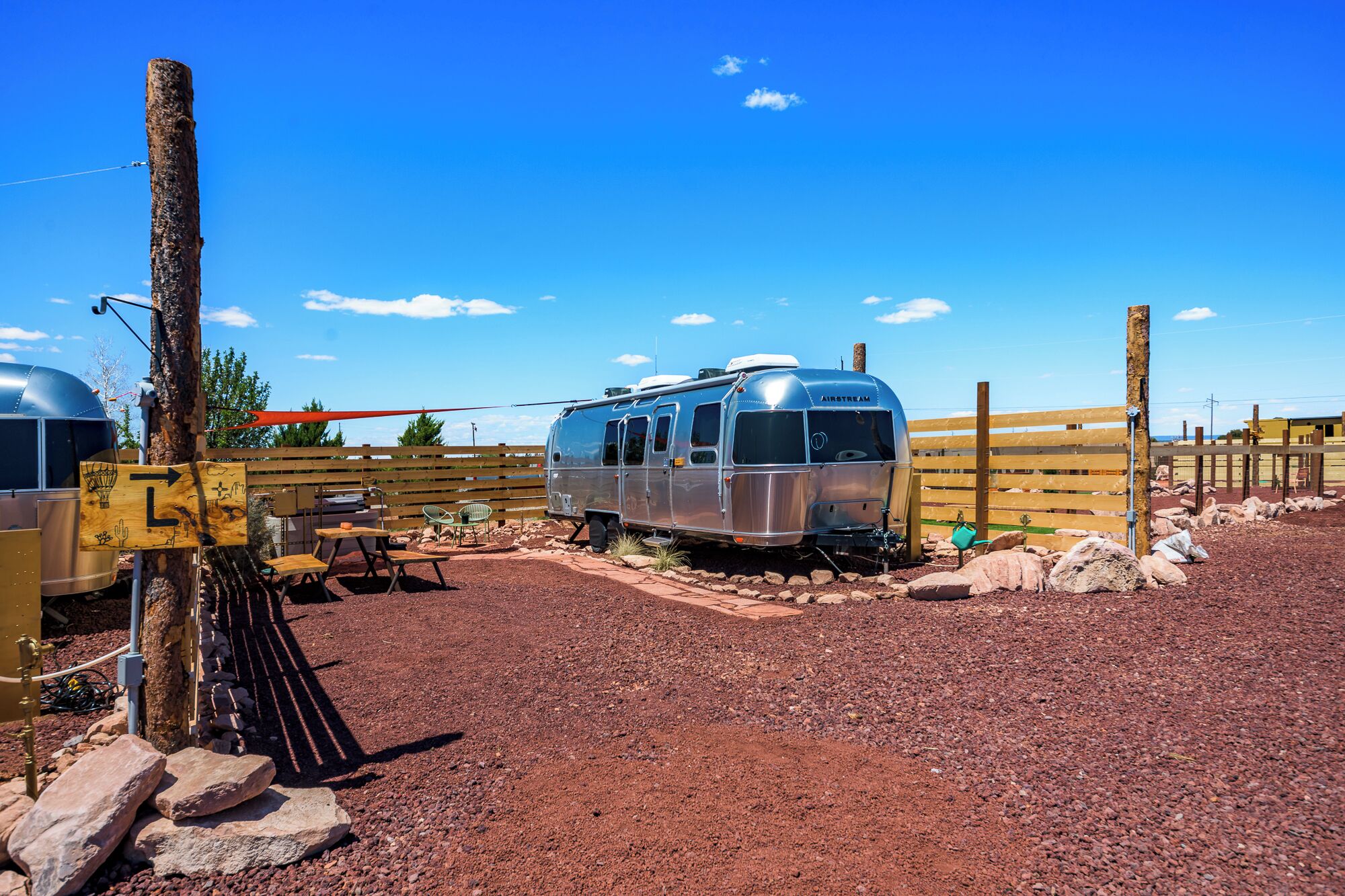 A vintage Airstream trailer parked in a gravel area with a wooden fence, picnic table, and a clear blue sky.