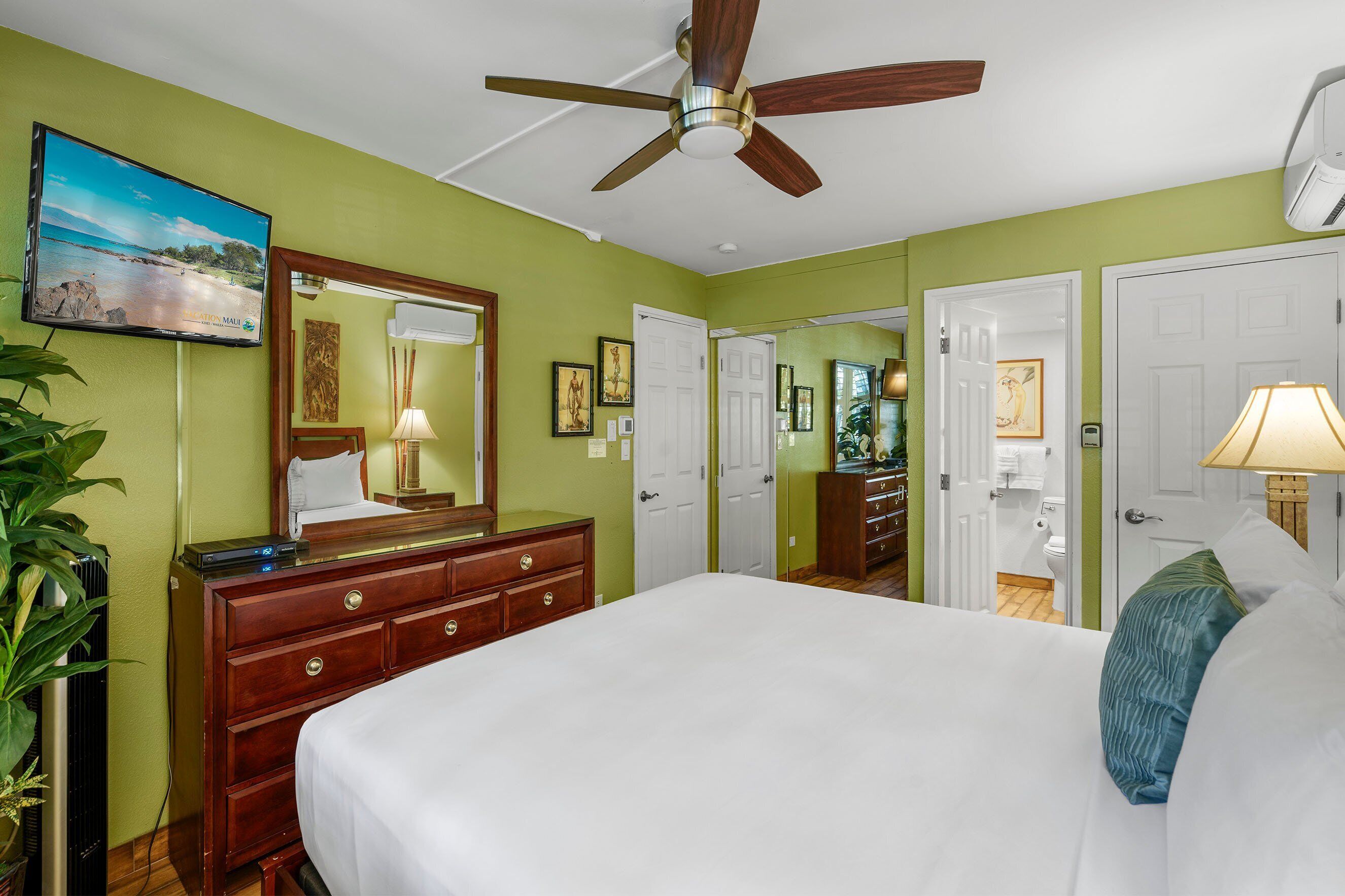 A hotel room with a bed, a dresser, a ceiling fan, a TV, and a door leading to a bathroom.