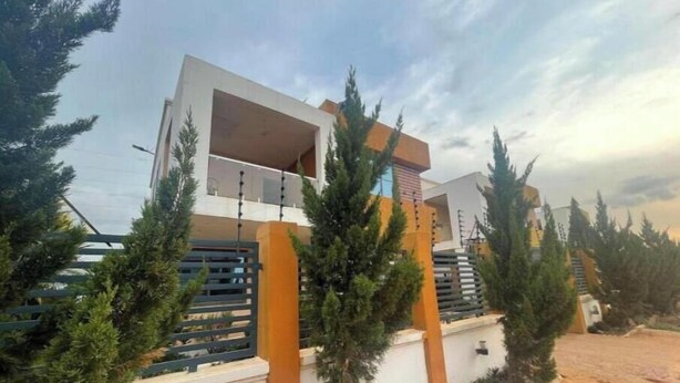 A modern house with a white facade and orange accents, surrounded by evergreen trees. - KUMASI's SECRET II (Juahaw)
