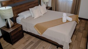 Cama bem arrumada com lençóis brancos e manta marrom, duas luminárias de cabeceira e cabeceira de madeira.