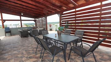 An outdoor patio with a stone-tiled floor, a wooden pergola, a table with chairs, and a grill.