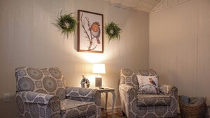 Two patterned armchairs, a small table with a lamp, and a wicker basket against a wall with a framed bird painting and greenery.