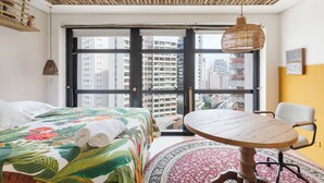 A hotel room with a large window, a bed with a floral bedspread, a round wooden table, and a chair. - Urbano Duplex by WECARE Hosting (São Paulo)