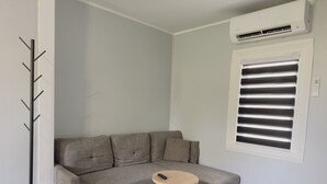 A corner room with a grey sectional sofa, a small round table, a coat rack, and a window with blinds. - Zen Den Getaway, your peaceful retreat near Cleveland’s vibrant AsiaTown. (Cleveland)