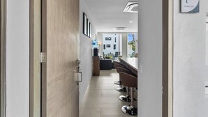 Un couloir d’hôtel moderne avec une porte en bois, un coin bar avec des tabourets et un distributeur d’eau visible.
