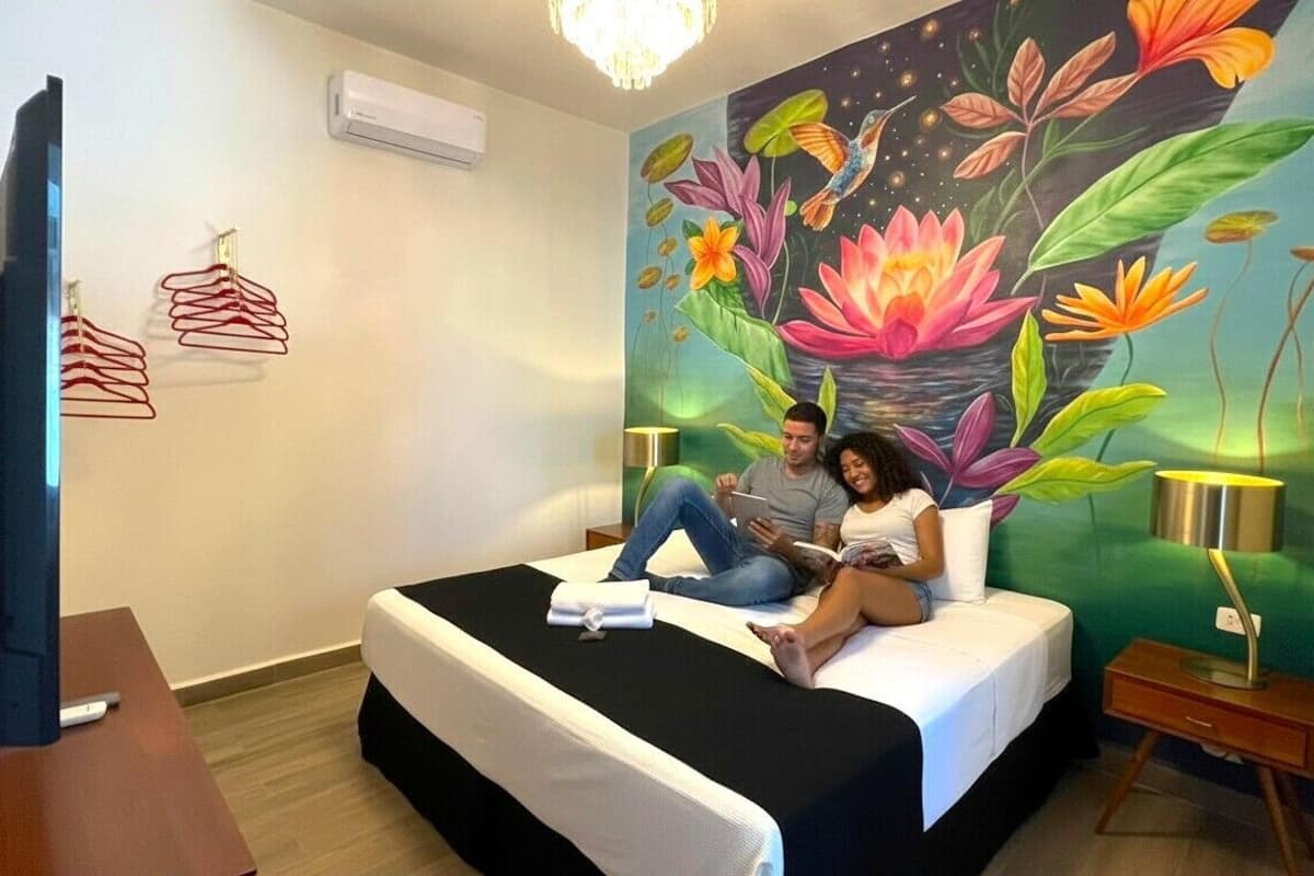 A bedroom with a bed, a bedside table with a lamp, a wall-mounted air conditioner, and a wall with a colorful mural.