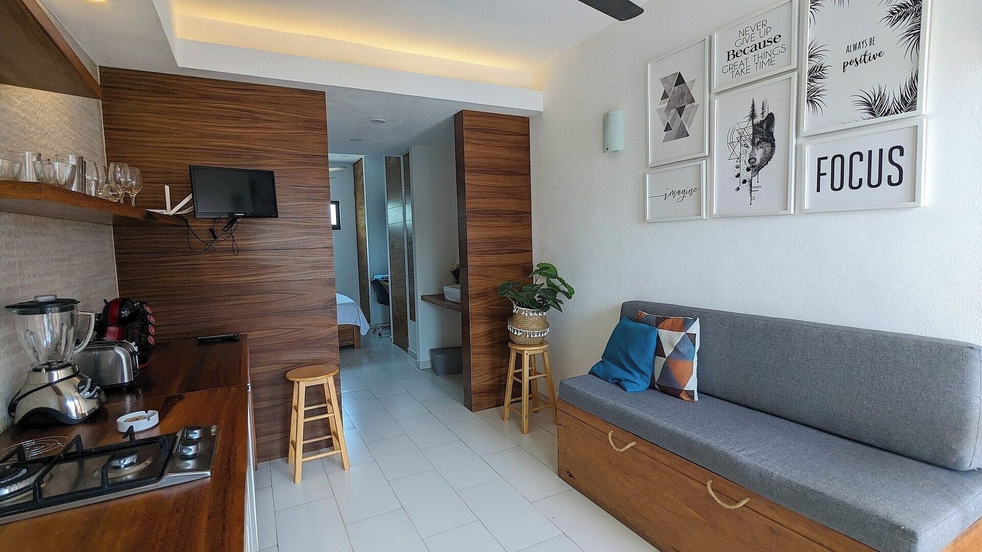 A modern kitchen with a built-in stove, a dining area with a grey sofa and a small table, and a living area with a TV and motivational wall art.