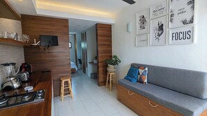 Una cocina moderna con estufa integrada, una zona de comedor con sofá gris y una mesita, y una sala de estar con televisor y decoración mural inspiradora.