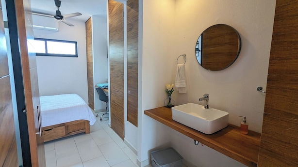 Habitación de hotel compacta con cama, pequeño escritorio, zona de ducha y baño con lavabo y espejo.