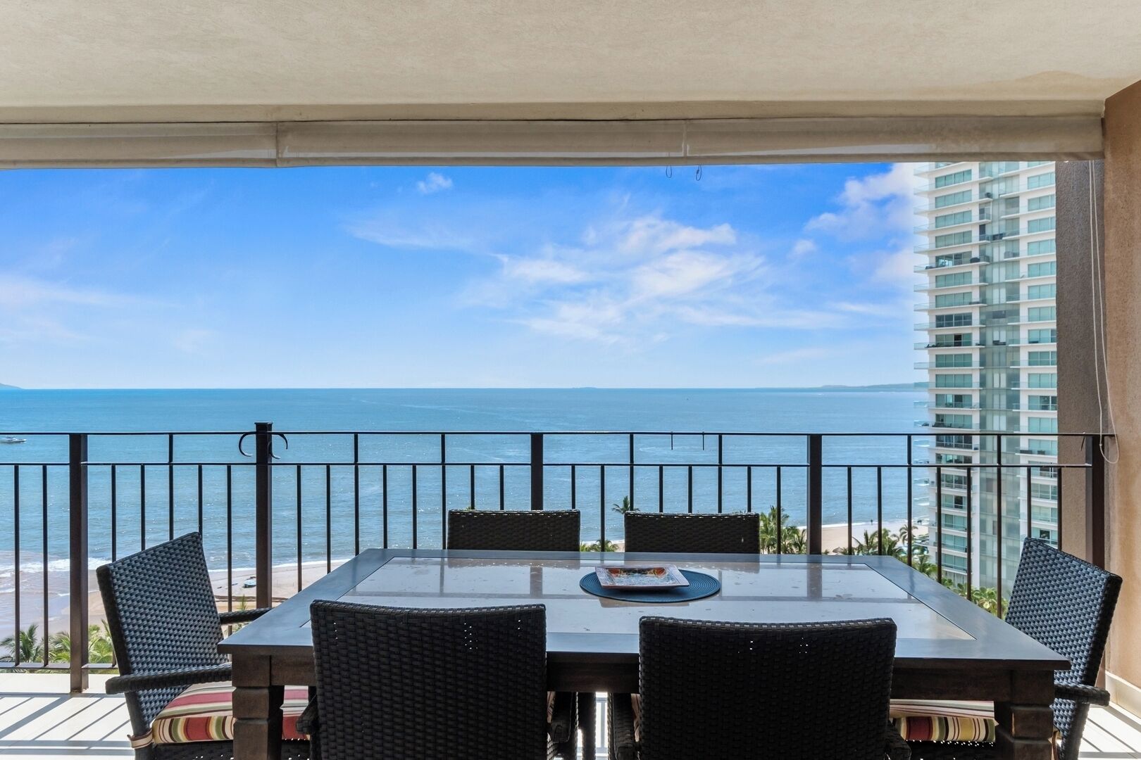 A balcony with a glass table and wicker chairs, overlooking a beach and ocean.