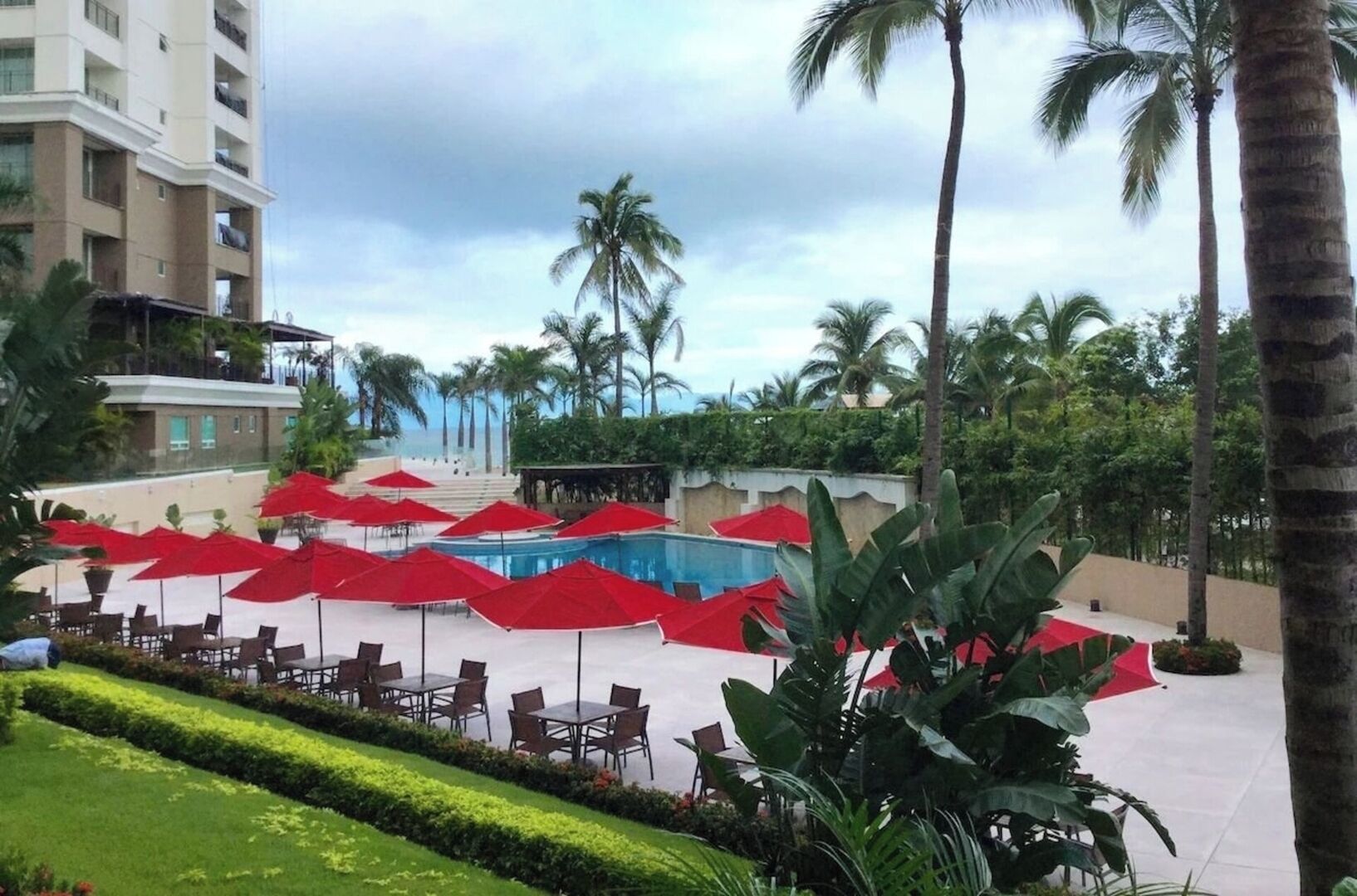 Ein Hotelpoolbereich mit roten Sonnenschirmen, Palmen und freiem Blick auf den Ozean.
