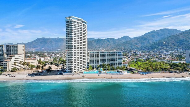 A coastal cityscape with a prominent high-rise hotel, beachfront, and mountainous backdrop. - Magnificent view from the floor 26 (Puerto Vallarta)
