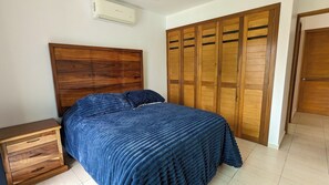 Ein Schlafzimmer mit Holz-Kopfteil, einem Bett mit blauer Bettdecke, einem Nachttisch aus Holz und einem Kleiderschrank mit mehreren Türen.