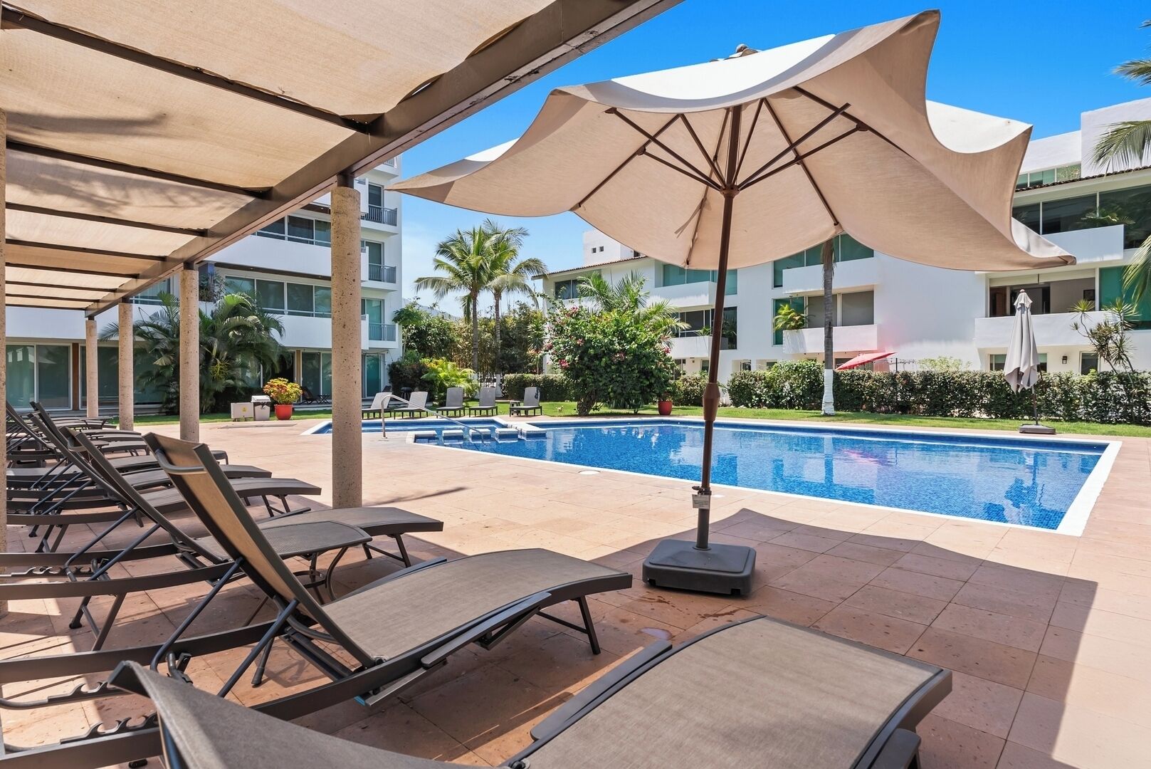 Uma área à beira da piscina com espreguiçadeiras e guarda-sóis, uma piscina azul cristalina e um edifício com varandas ao fundo.