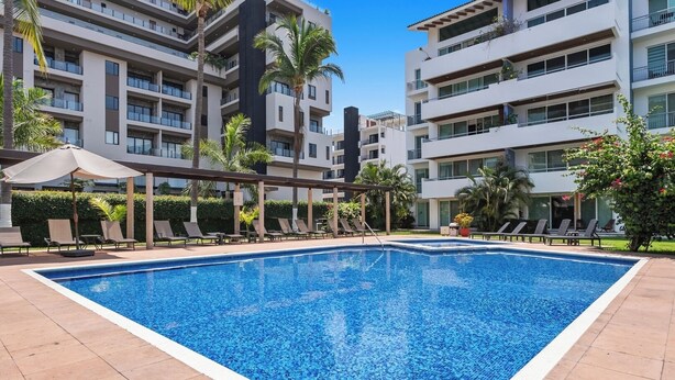 A swimming pool with lounge chairs, surrounded by multi-story buildings with balconies and palm trees. - Cozy Studio, great amenities in Versalles-Fluvial (Puerto Vallarta)