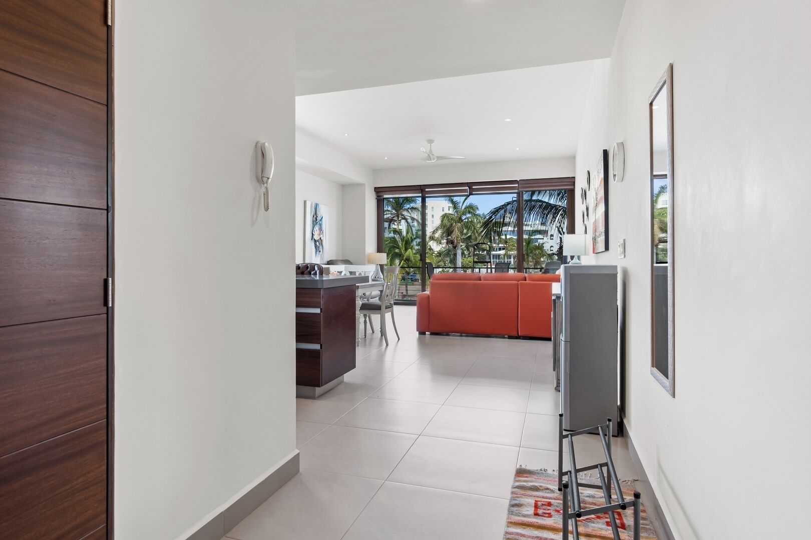 A modern kitchen with a dark wood cabinet, a dining area with a red sofa, and a view of palm trees through large windows.