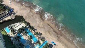 Vista aérea de una playa con aguas turquesas cristalinas, una orilla arenosa y una zona de resortes con piscinas y palmeras.