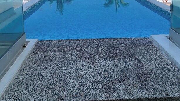 Una piscina infinita de color azul cristalino con borde de textura de guijarros, bordeada por palmeras, que se extiende hasta una playa.