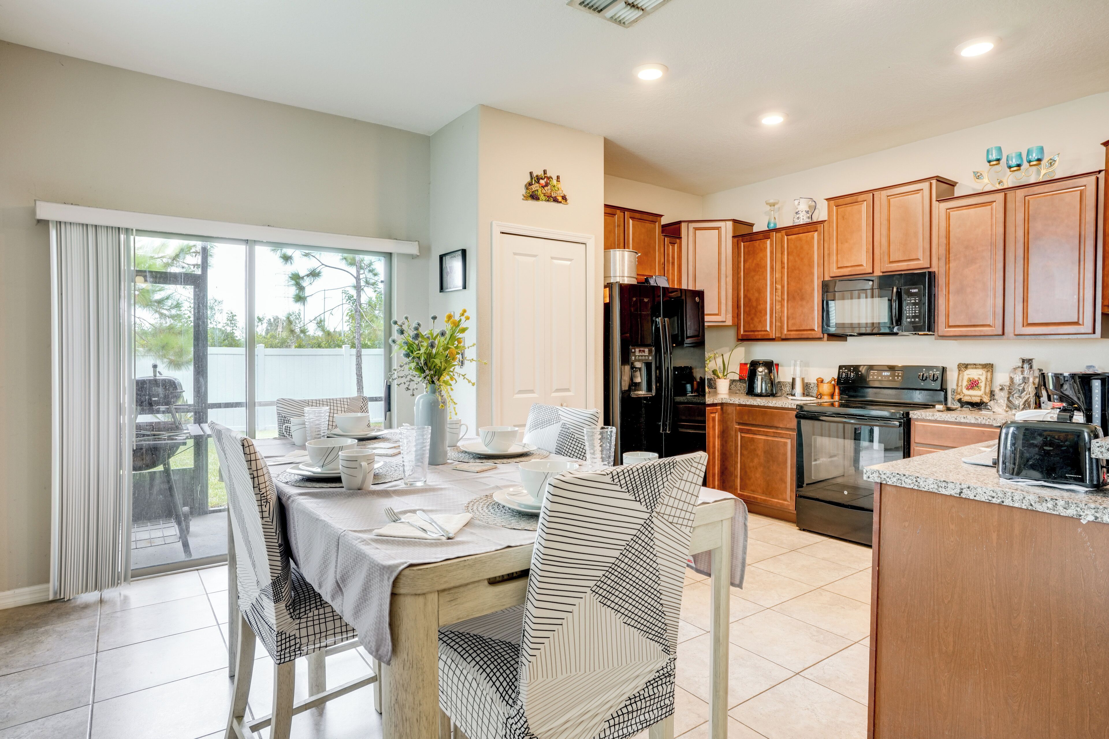 A modern kitchen with wooden cabinets, a refrigerator, microwave, and stove. There is a dining area with a table set for two, chairs, and a vase with flowers.