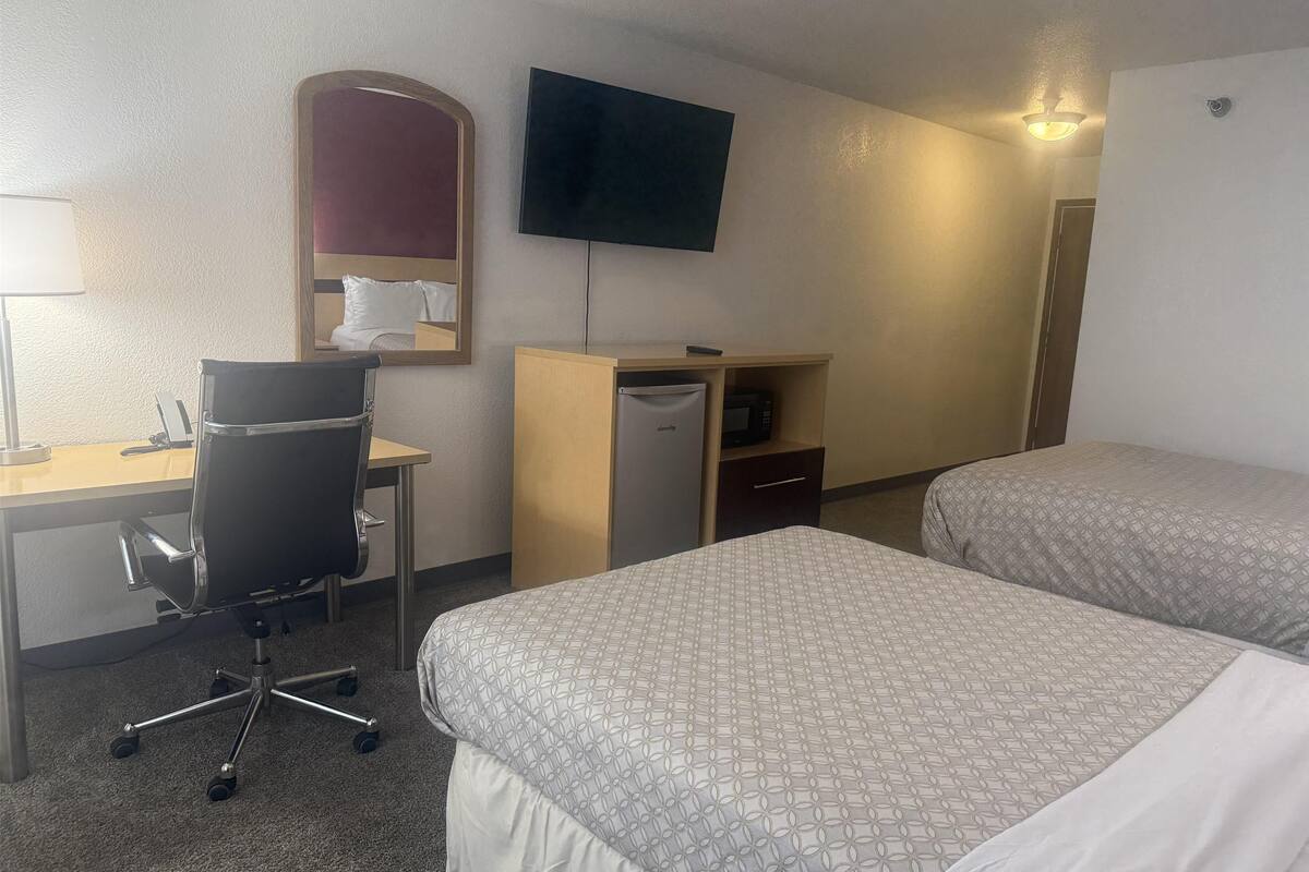 Ein Hotelzimmer mit zwei Betten, einem Schreibtisch, einem Stuhl, einem Kühlschrank, einer Mikrowelle, einem Fernseher und einem Spiegel.