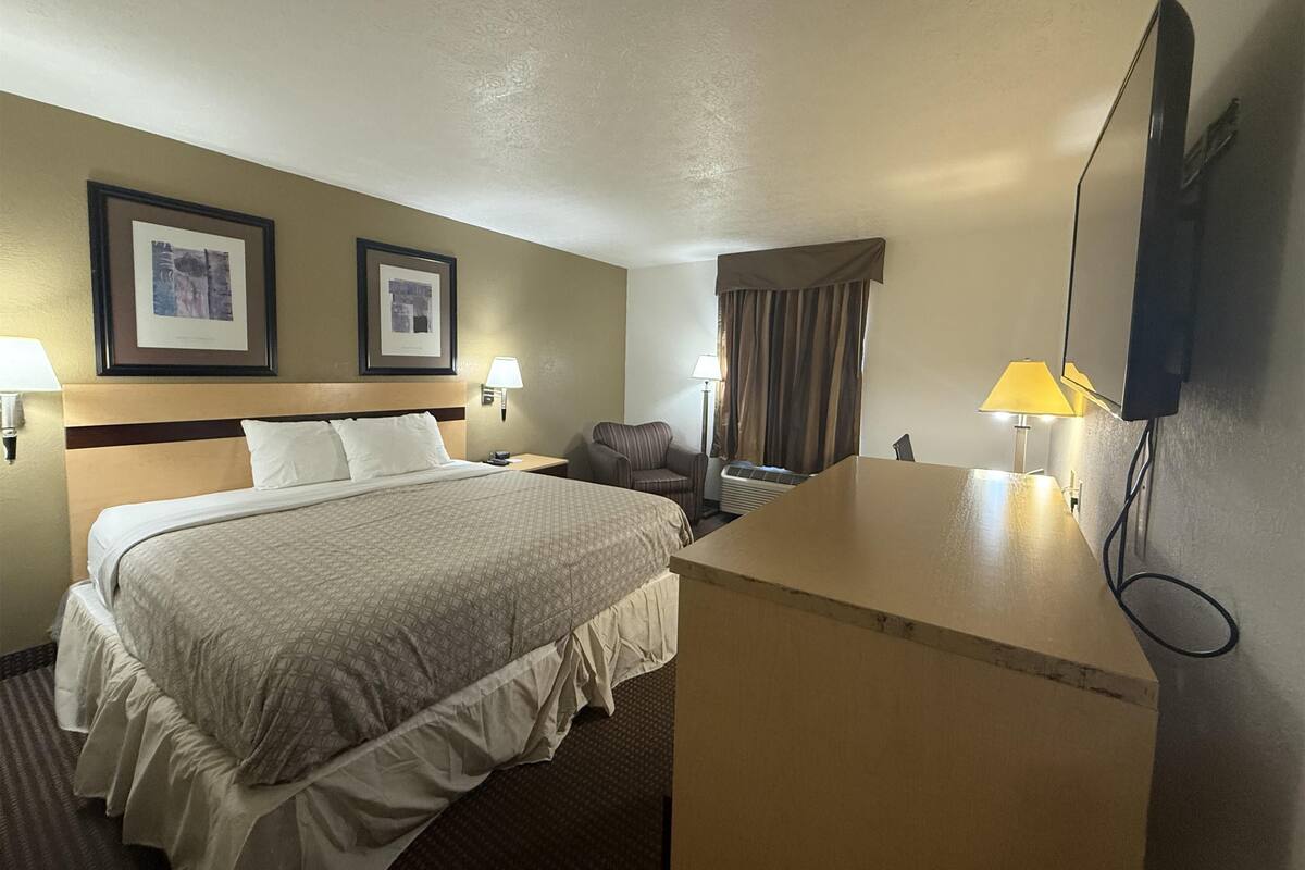 Ein Hotelzimmer mit einem Bett, zwei Nachttischlampen, einem Schreibtisch mit Lampe, einem Sessel, einem Fernseher und zwei gerahmten Bildern an der Wand.