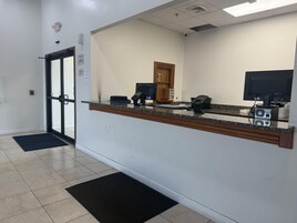 A reception desk with a computer, phone, and a monitor. There is a door with a glass panel and a fire alarm on the wall.