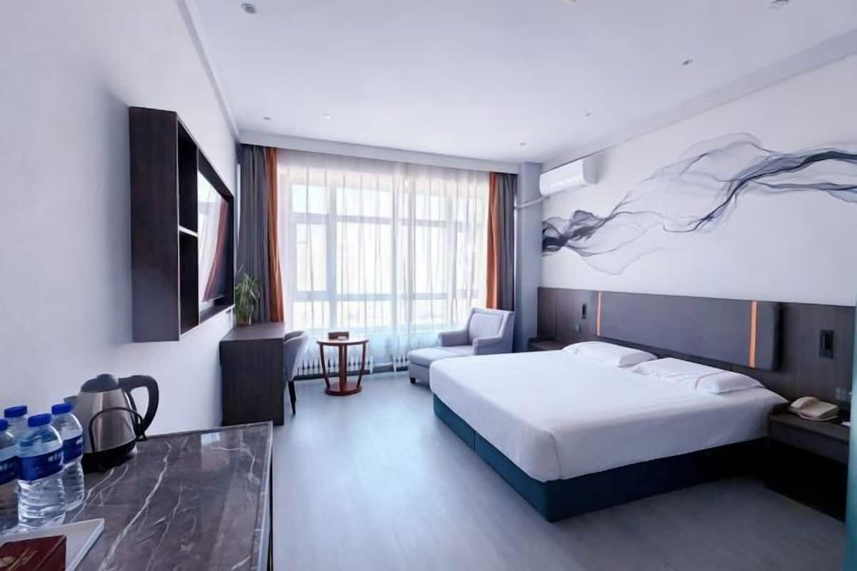 A modern hotel room with a large bed, a desk with a chair, a small table, and a large window with curtains.
