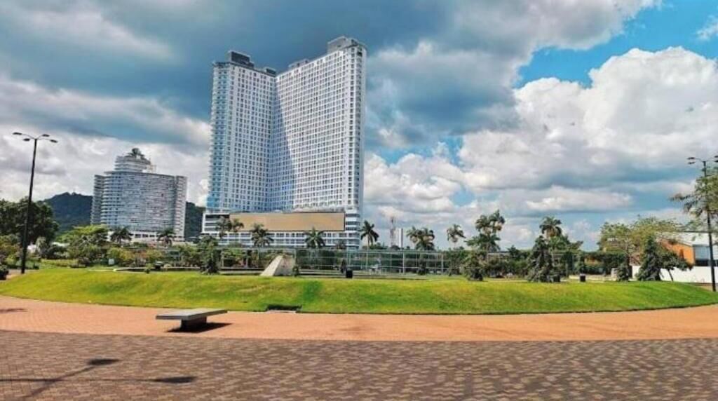 A high-rise hotel with a modern design, surrounded by a landscaped park area with a paved walkway and benches.