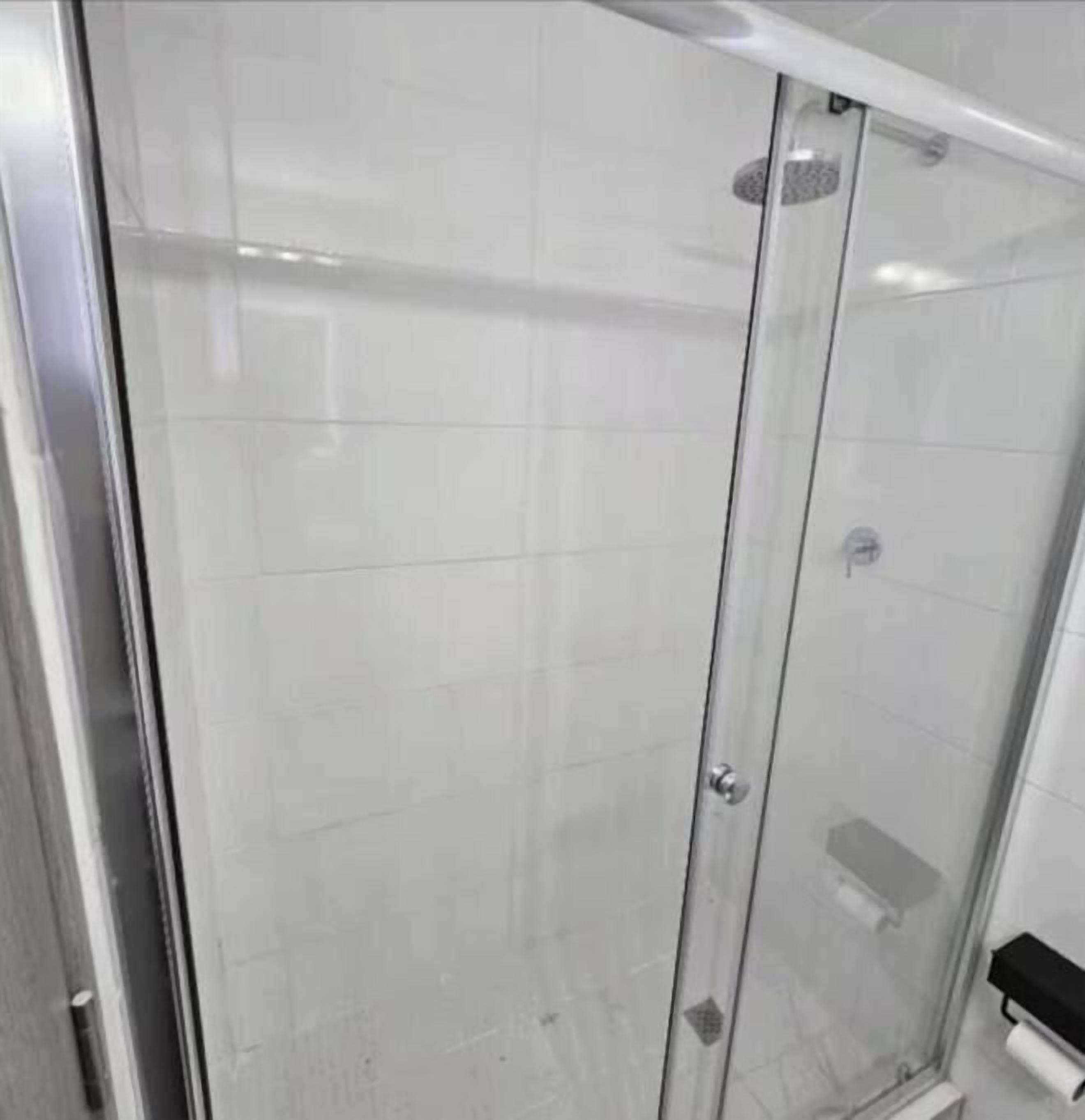 A glass-enclosed shower stall with a tiled wall, a door with a handle, and a small shelf with a roll of toilet paper.