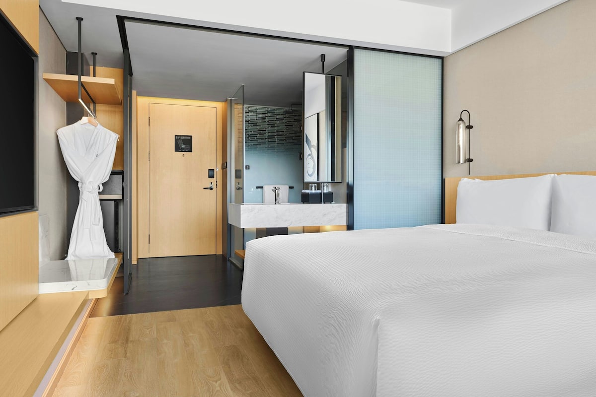 A modern hotel room with a large bed, a wooden floor, a bathroom with a mirror and sink, and a robe hanging on a rack.