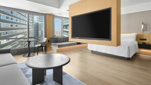 Habitación de hotel moderna con un televisor grande, asientos cómodos, un balcón con vistas a la ciudad y un ventanal amplio.