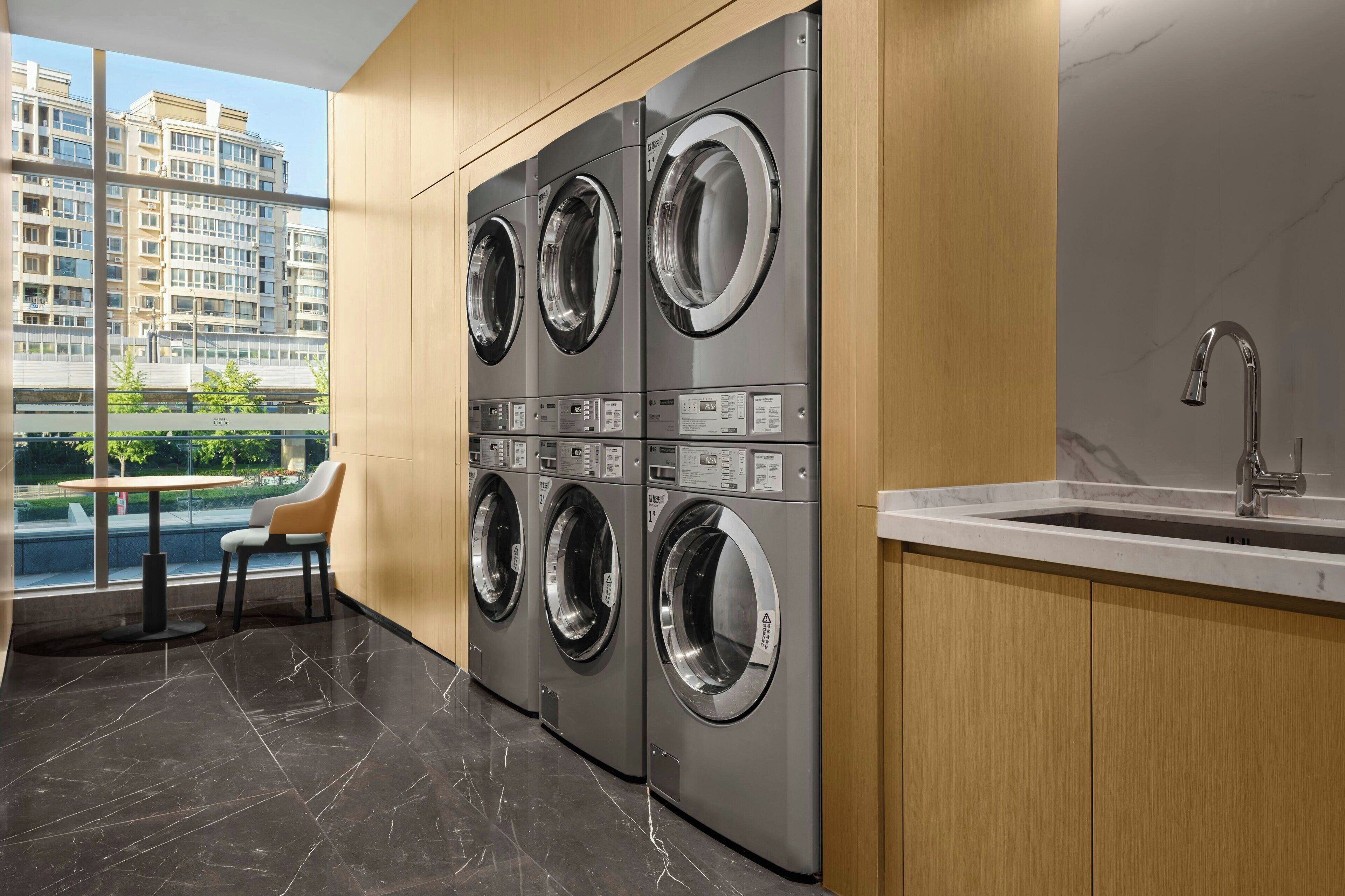 A modern laundry room with four stacked front-loading washing machines, a sink, and a view of a residential building through large windows.
