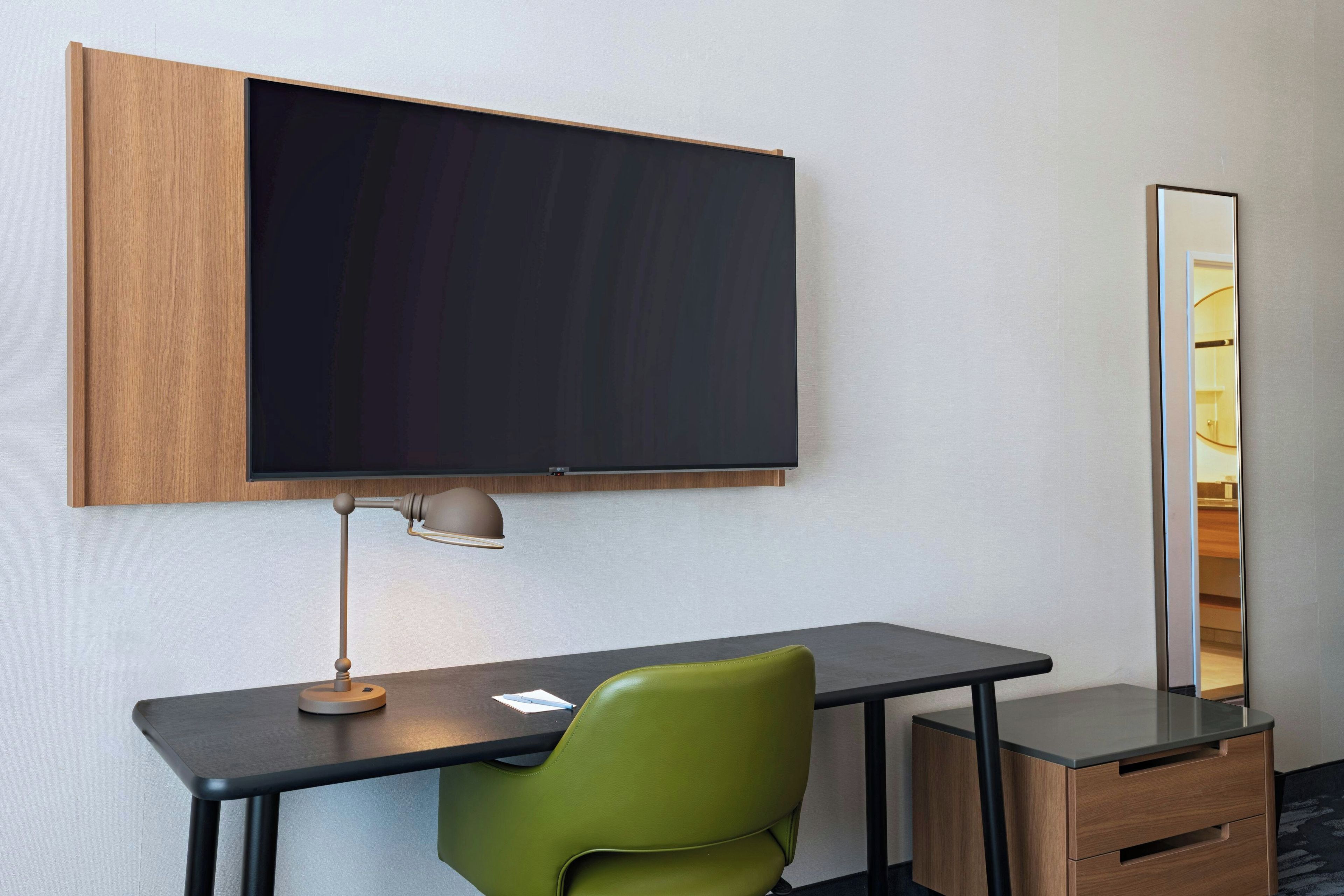 A hotel room with a desk, a green chair, a flat-screen TV mounted on the wall, a desk lamp, and a mirror.
